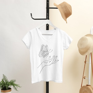 May include: White V-neck t-shirt with a black line drawing of a butterfly flying towards an outstretched hand.