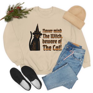 Puede incluir: Una sudadera beige con un gato negro con un sombrero de bruja y el texto "Never mind The Witch, beware of The Cat!"