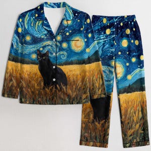Op de afbeelding: Een tweedelige pyjamaset met een zwarte kat in een tarweveld onder een sterrenhemel. De top is een overhemd met knopen en de broek heeft een bijpassend ontwerp. Het kleurenpalet omvat blauw, geel en zwart.