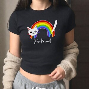 Puede incluir: Una camiseta negra con un gato blanco y un arcoíris. El gato está enroscado alrededor del arcoíris y el texto "Be Proud" está impreso debajo del gato.