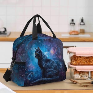Könnte beinhalten: Schwarze Katzen-Lunchtasche mit Galaxie-Design. Die Tasche zeigt eine schwarze Katze mit gelben Augen vor dem Hintergrund eines blauen und violetten Nebels. Sie hat einen schwarzen Schultergurt und Griffe.