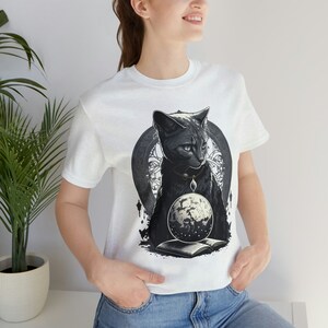 May include: White t-shirt featuring a black cat design with blue eyes, a necklace, and a moon-like orb. The cat is set against a circular, ornate background. The design includes an open book at the bottom.
