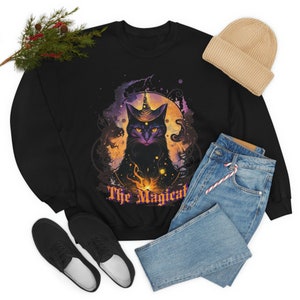 May include: Black sweatshirt featuring a mystical cat design with a witch's hat and the text "The Magical". The graphic includes a purple and orange celestial background. A beige beanie, blue jeans, and black shoes are also visible.