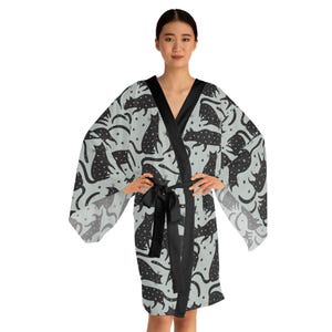 Op de afbeelding: Lichtgrijze kimono-achtige badjas met een zwart kattenpatroon en een zwarte ceintuur. De badjas heeft een V-hals en wijde mouwen. De katten staan in verschillende poses, met sterren verspreid.