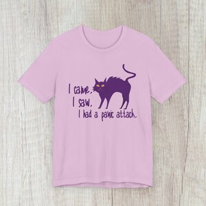 May include: A light pink t-shirt featuring a purple cat silhouette with yellow eyes. The text "I came. I saw. I had a panic attack." is printed in purple below the cat graphic. The shirt is made of soft material.