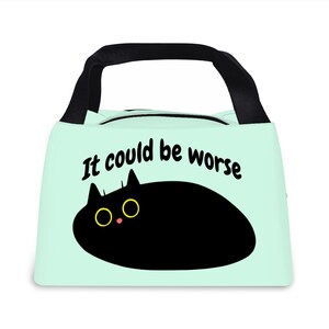 Op de afbeelding: Een lichtblauwe lunchtas met een zwarte handgreep en ritssluiting. De tas heeft een cartoon zwarte kat met gele ogen en de zin "It could be worse" in zwarte tekst.