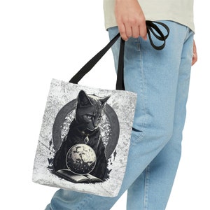 May include: A black cat with a silver necklace and a celestial globe sits on a book in front of a circular design. The cat is on a white tote bag with a black handle.