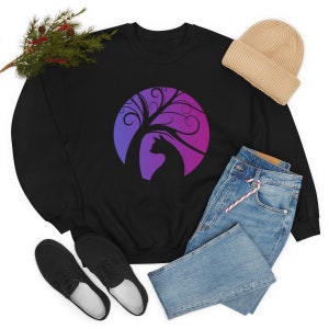 May include: Black sweatshirt with a cat and tree design in purple and blue. A beige beanie and blue jeans are next to the sweatshirt. Black shoes are at the bottom left. The design is set against a circular background.