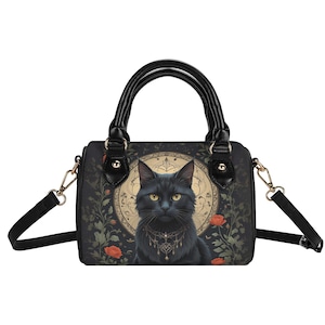 May include: Black handbag featuring a detailed illustration of a black cat with yellow eyes, adorned with a decorative necklace. The bag has a black handle and a detachable shoulder strap. The background includes floral and ornamental elements.