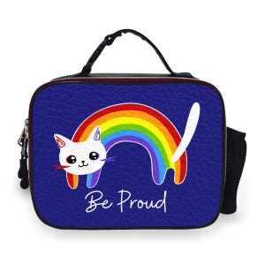 May include: A blue lunch bag featuring a white cat with a rainbow and the text "Be Proud". The cat has a red tongue and is outlined in black. The rainbow arches over the cat. The bag has a black handle and a side mesh pocket.