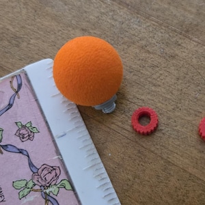 May include: A bright orange spherical foam ball with a small grey plastic base, positioned next to a white ruler and two small red plastic circular rings with ridged edges. The ruler shows measurements in centimetres, with the ball measuring 2.5 cm.