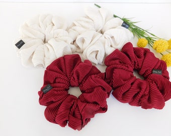 May include: A selection of pleated fabric scrunchies in cream and burgundy. Each scrunchie has a small black tag. The scrunchies are displayed on a white surface with greenery and yellow flowers.
