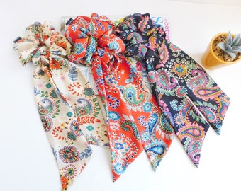 May include: Four colorful scrunchies with long, patterned fabric tails. The scrunchies feature paisley and floral designs in cream, red, navy, and orange. The fabric tails add a decorative element to these hair accessories.