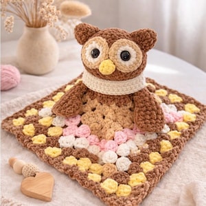 Könnte beinhalten: Gehäkelte Eule mit großen Augen und gelbem Schnabel, trägt einen cremefarbenen Schal. Die Eule sitzt auf einer quadratischen Decke mit einem Granny-Square-Muster in Braun, Rosa, Gelb und Weiß. Ein herzförmiger Beißring aus Holz liegt daneben. Die Decke misst ca. 30 cm x 30 cm.