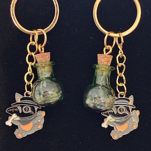 May include: A pair of gold hoop earrings. Each earring has a small green glass bottle with a cork stopper, a gold chain, and a charm of a cartoon dog wearing a hat and holding a bone. The dog charm is grey, black, and orange.