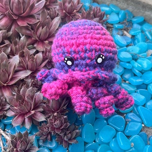 May include: A crocheted octopus toy with pink and purple stripes. The octopus has large black eyes and is sitting on a bed of blue pebbles.