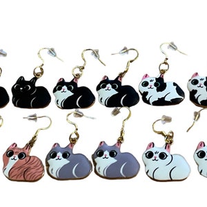 May include: A set of 8 cat-shaped dangle earrings. The earrings are made of metal and have a gold finish. The cats are painted in various colors, including black, white, gray, and orange.