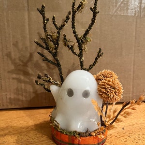 May include: A white ghost figurine with black eyes sits on a plaid base surrounded by dried flowers and branches.