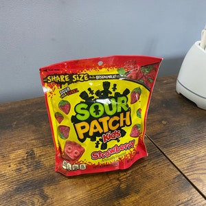 Könnte beinhalten: Ein wiederverschließbarer Beutel mit Sour Patch Kids Erdbeer-Bonbons in Rot, Gelb und Grün. Der Beutel zeigt eine Comicfigur und den Text "Sour Patch Kids Strawberry" und "Artificially Flavored".