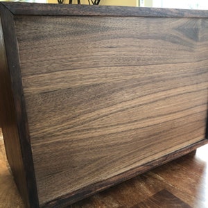 Puede incluir: Una caja de madera marr&oacute;n oscuro con un acabado liso y pulido. La caja tiene una forma rectangular simple y un borde ligeramente elevado alrededor de la parte superior.