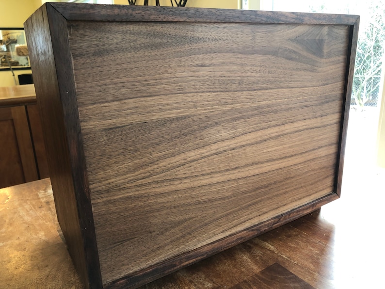 Puede incluir: Una caja de madera marr&oacute;n oscuro con un acabado liso y pulido. La caja tiene una forma rectangular simple y un borde ligeramente elevado alrededor de la parte superior.