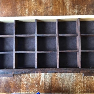 Puede incluir: Una caja de madera con un forro de fieltro marr&oacute;n y 16 compartimentos individuales. La caja est&aacute; hecha de madera clara y oscura.