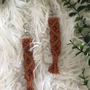 May include: Two brown macrame keychains with silver key rings and tassels. The keychains are hanging on a white fluffy surface.