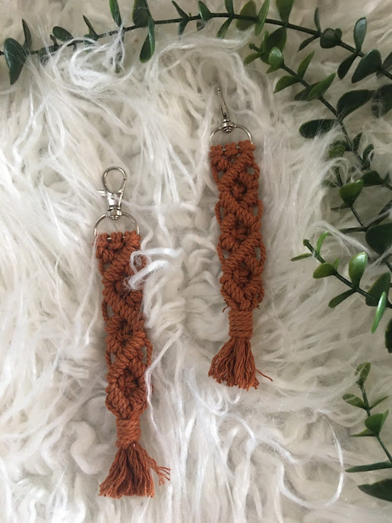 May include: Two brown macrame keychains with silver key rings and tassels. The keychains are hanging on a white fluffy surface.