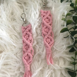 May include: Two pink macrame keychains with silver key rings and tassels. The keychains are made with a knotting technique and have a delicate, feminine look.