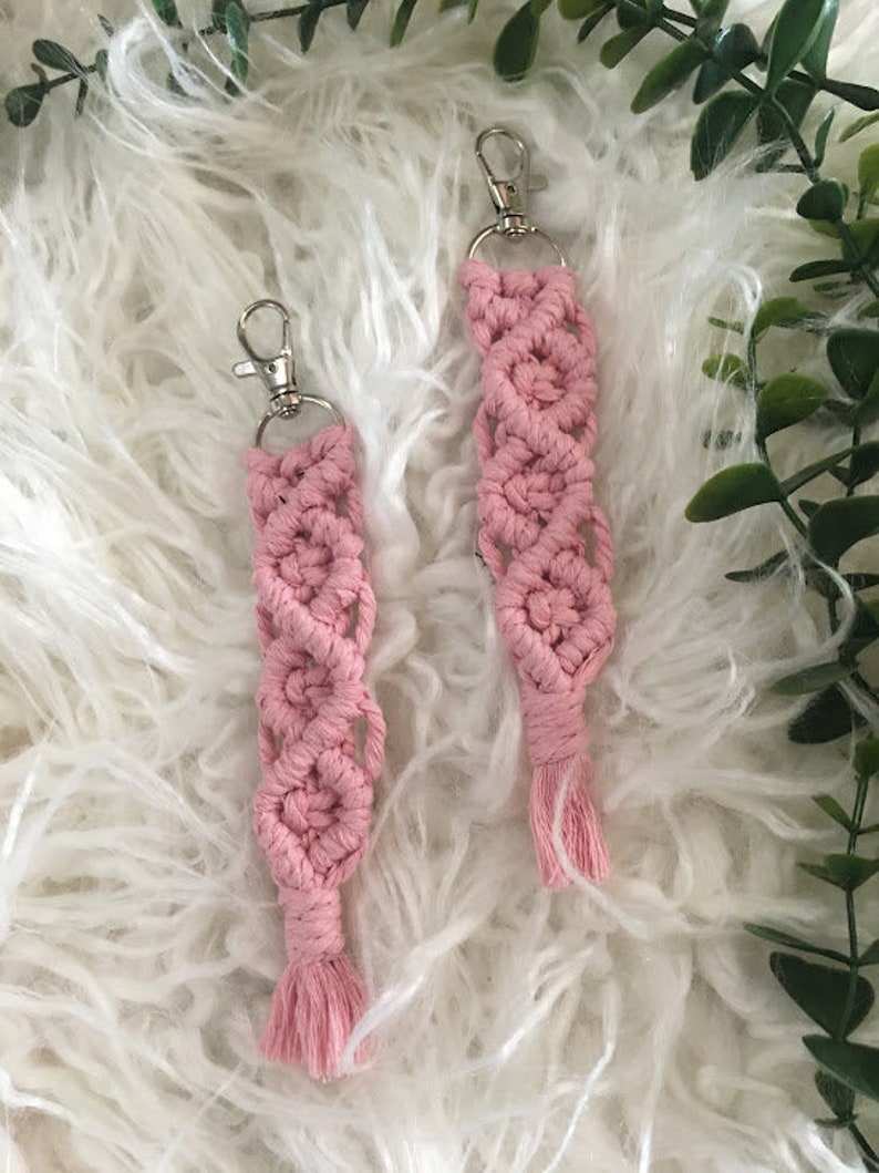 May include: Two pink macrame keychains with silver key rings and tassels. The keychains are made with a knotting technique and have a delicate, feminine look.