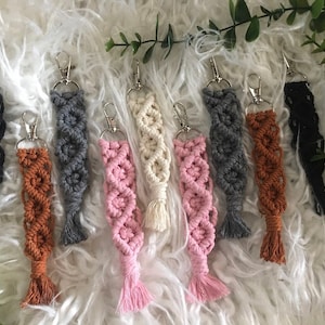 May include: A set of eight colorful macrame keychains with silver key rings. The keychains are made with a variety of colors, including black, brown, gray, pink, and white.