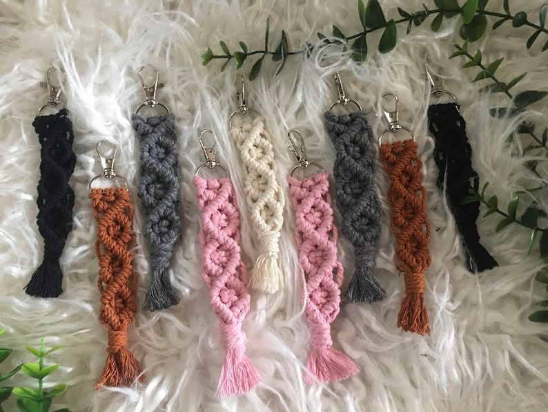 May include: A set of eight colorful macrame keychains with silver key rings. The keychains are made with a variety of colors, including black, brown, gray, pink, and white.