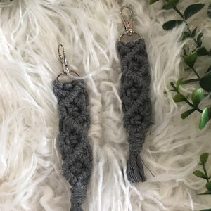May include: Two gray macrame keychains with silver key rings and tassels. The keychains are on a white fluffy surface.