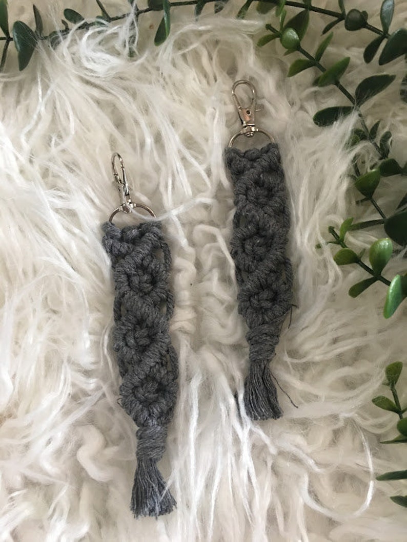 May include: Two gray macrame keychains with silver key rings and tassels. The keychains are on a white fluffy surface.