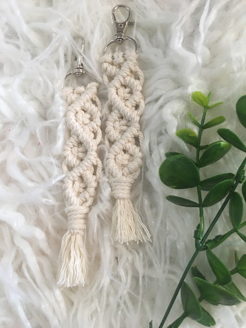 May include: Two white macrame keychains with silver key rings and tassels. The keychains are made with a knotting technique and have a geometric pattern.