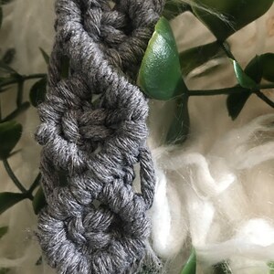 May include: A gray macrame cord with a repeating pattern of knots, creating a decorative design. The cord is partially visible against a white fuzzy background and green leaves.