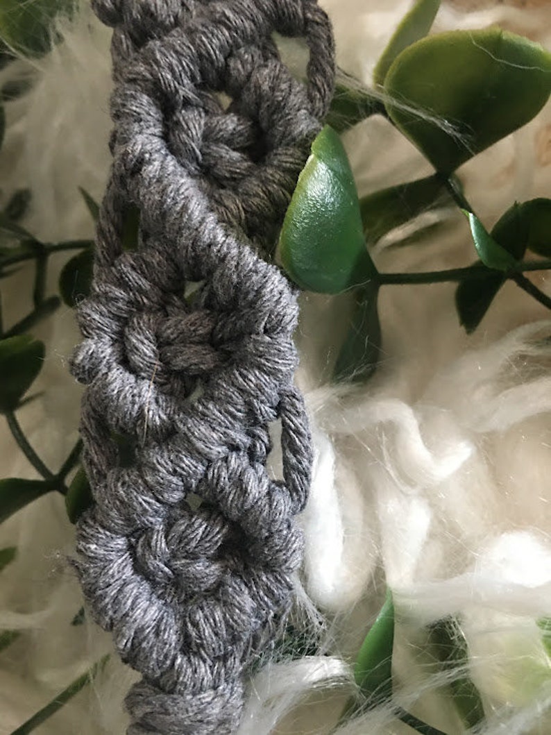 May include: A gray macrame cord with a repeating pattern of knots, creating a decorative design. The cord is partially visible against a white fuzzy background and green leaves.