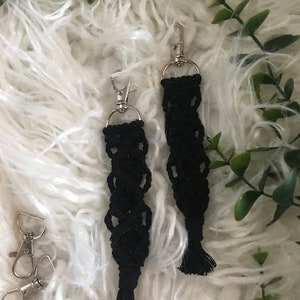 May include: Two black macrame keychains with silver key rings and tassels. The keychains are hanging on a white fluffy surface.