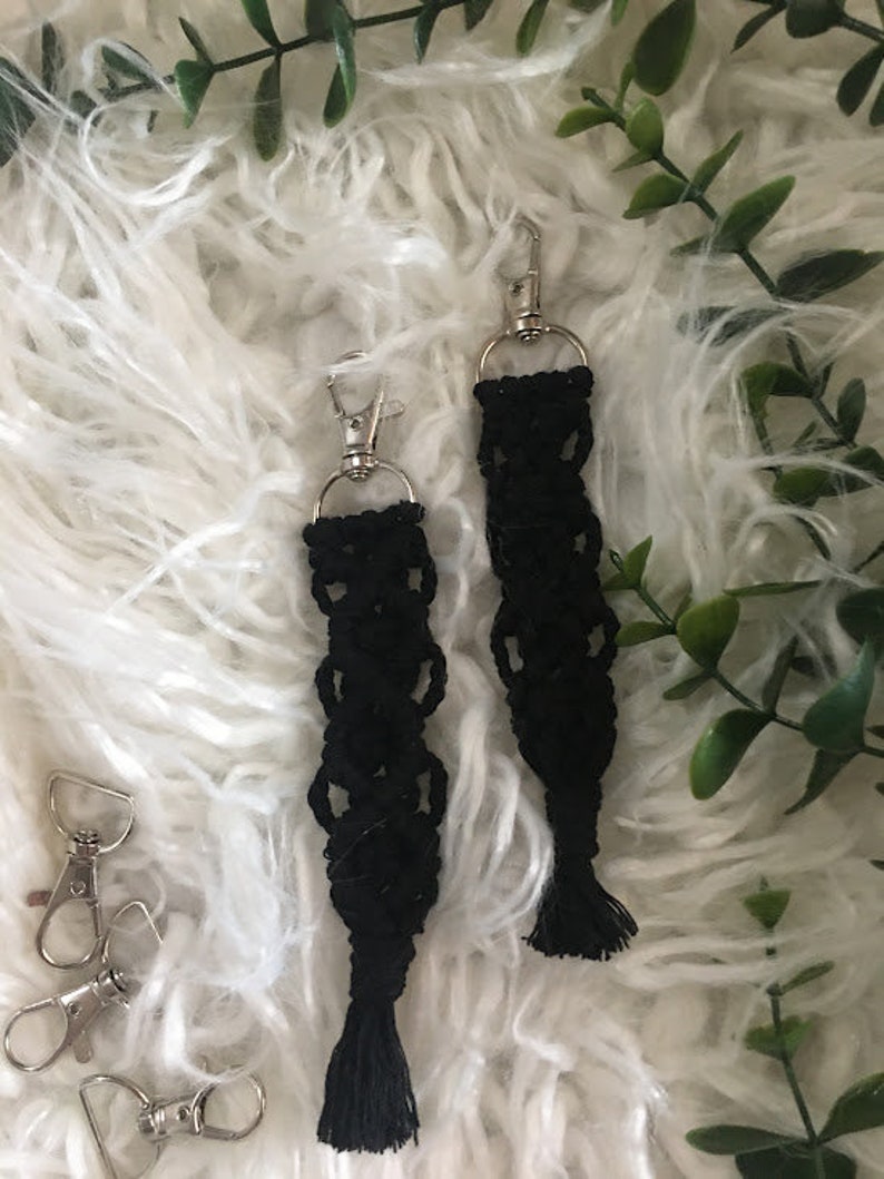 May include: Two black macrame keychains with silver key rings and tassels. The keychains are hanging on a white fluffy surface.