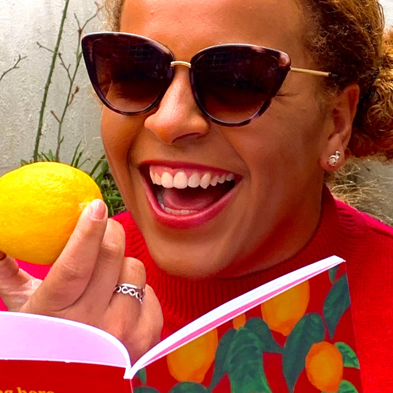 May include: A woman wearing sunglasses and a red sweater is holding a lemon and smiling. She is reading a book with a lemon illustration on the cover.