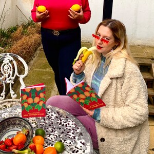 May include: Two women, one wearing a red sweater and the other a white fuzzy coat, are sitting at a table with fruit and a book that says "make every day nice enough to eat".