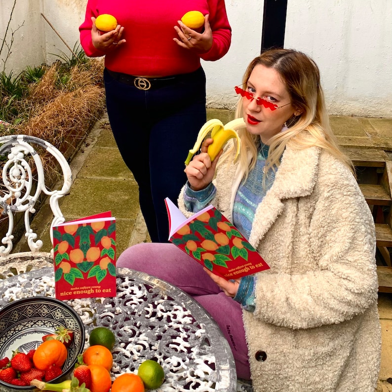 May include: Two women, one wearing a red sweater and the other a white fuzzy coat, are sitting at a table with fruit and a book that says "make every day nice enough to eat".