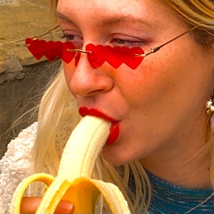 May include: A woman wearing red heart-shaped sunglasses bites into a yellow banana.