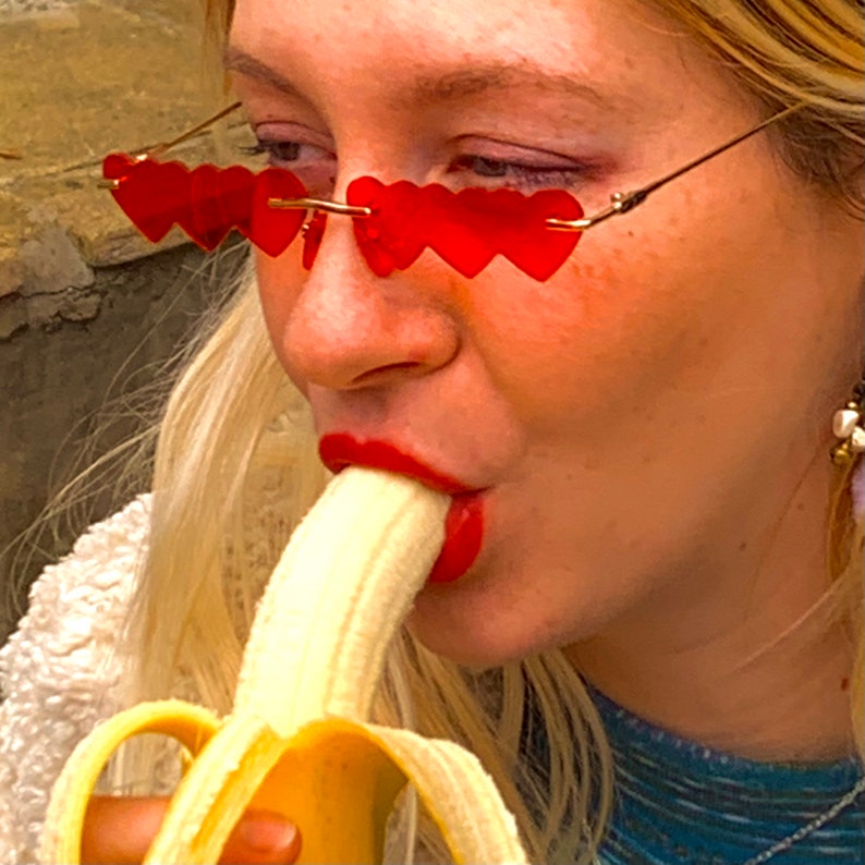May include: A woman wearing red heart-shaped sunglasses bites into a yellow banana.