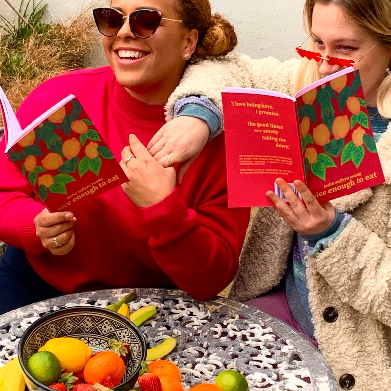 May include: Two friends are sitting at a table outside, reading books with lemon illustrations and the text "take silly you young, nice enough to eat". The books are red with a pink border. The table is made of metal and has a pattern of circles. There are lemons, limes, oranges, and strawberries in a bowl on the table.