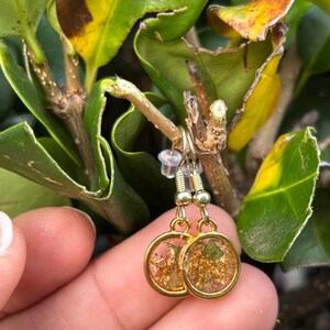 May include: Gold-colored dangle earrings with round, clear resin pendants. The pendants contain gold flakes and small green pieces. The earrings are hanging from a green leafy branch. The earrings are held by a person's hand.