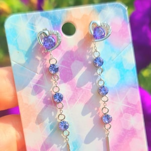 Peut inclure: Boucles d'oreilles pendantes avec un cœur orné de pierres violettes. Les boucles d'oreilles sont en métal argenté et comportent une barre dorée en bas. Présentées sur une carte aux couleurs vives.