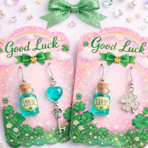 Può includere: Due paia di orecchini su biglietti rosa con la scritta "Good Luck". Un paio presenta una piccola bottiglia con il testo "Liquid Luck", l'altro un cuore e un portachiavi, e un quadrifoglio. Un fiocco verde è in alto.