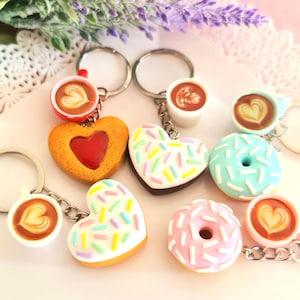 May include: Six keychains featuring different food and drink designs. The keychains include a heart-shaped biscuit with a red jelly centre, a heart-shaped doughnut with white icing and colourful sprinkles, a blue doughnut with white icing and sprinkles, a coffee cup with latte art, and a heart-shaped doughnut with pink icing and sprinkles.
