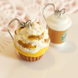 Puede incluir: Un par de pendientes novedosos con un cupcake y una taza de café. El cupcake tiene glaseado blanco con salsa de caramelo y una base amarilla. La taza de café es marrón con tapa blanca y un logotipo verde.